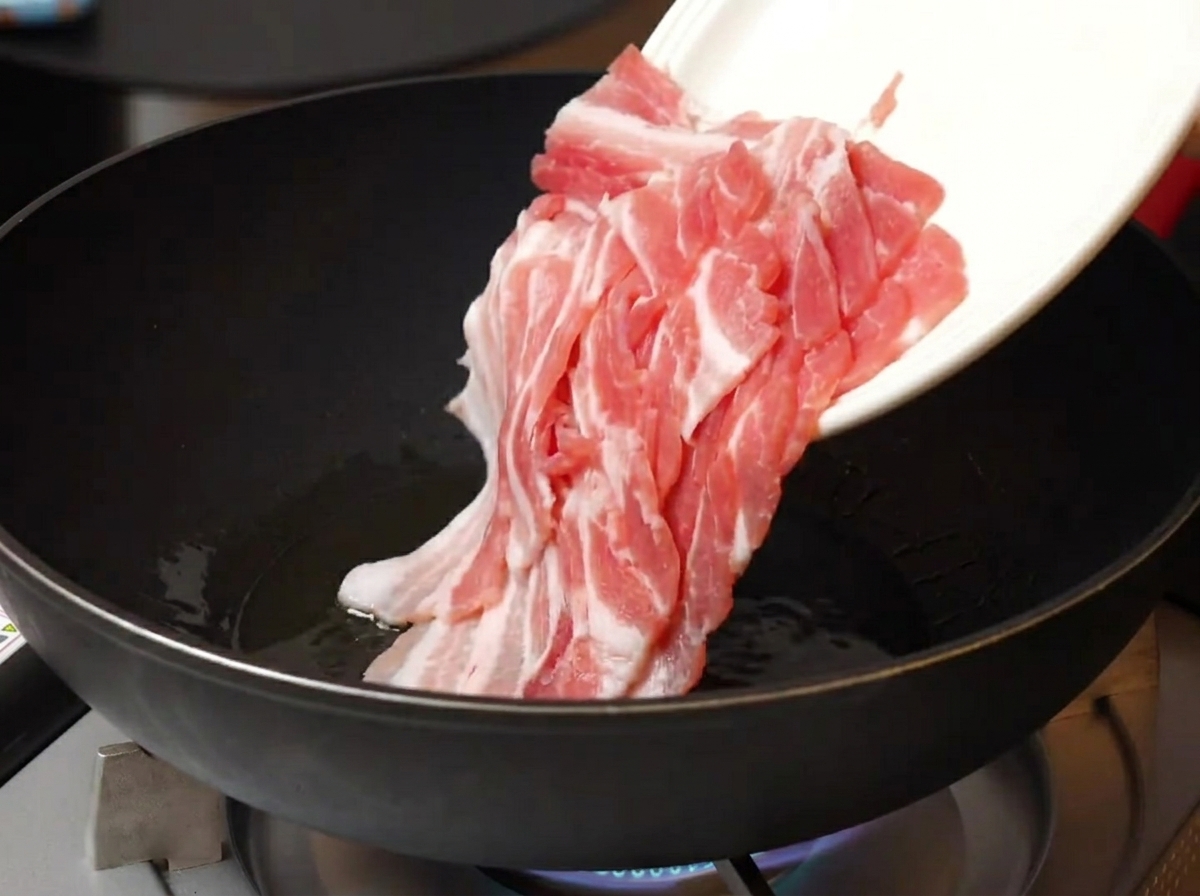 Dünn geschnittener roher Schweinebauch wird von einem weißen Teller in eine heiße, geölte Pfanne gegeben.