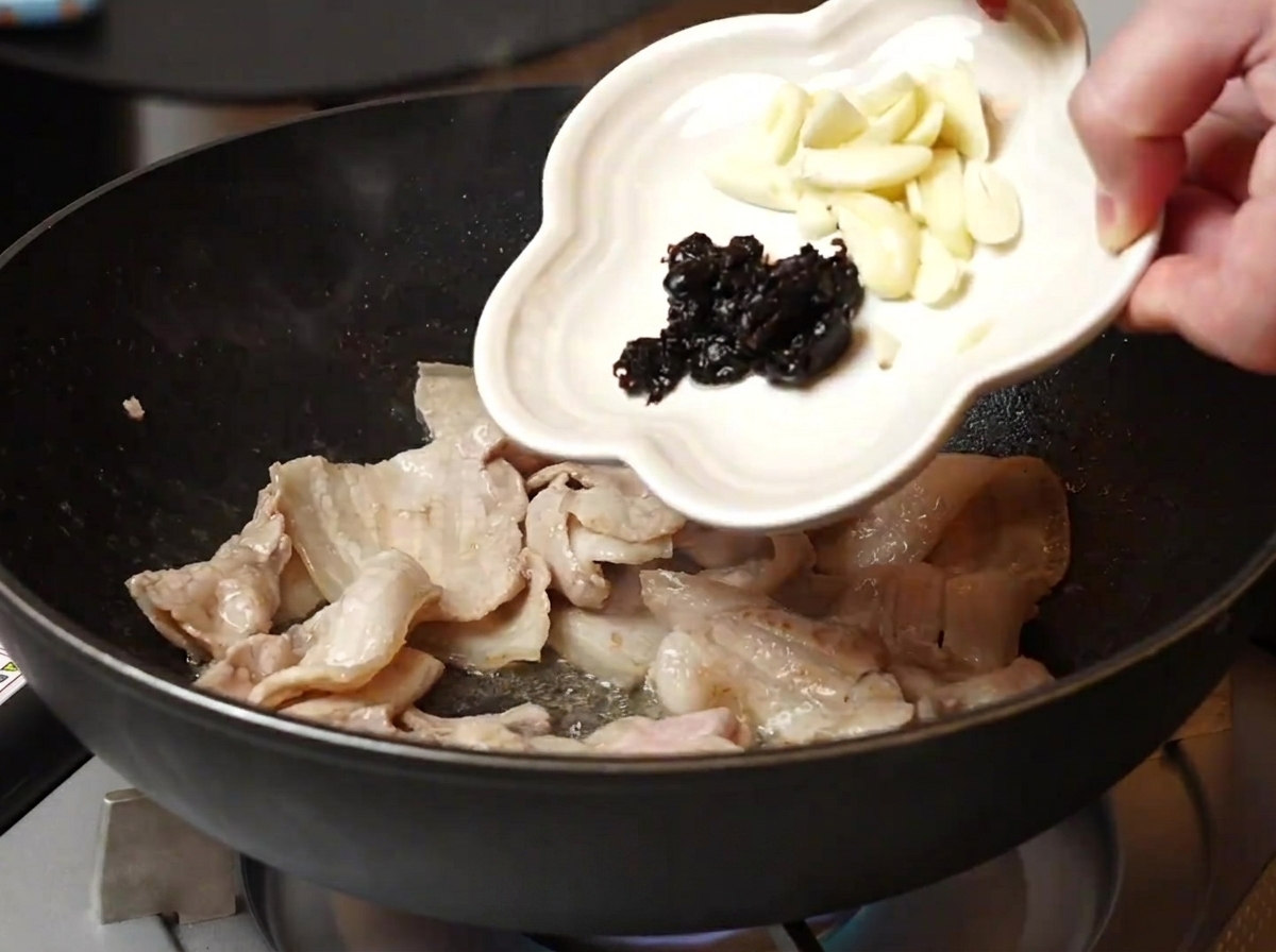 Knoblauchscheiben und fermentierte schwarze Bohnen werden aus einer kleinen weißen Schale in eine Pfanne mit gebratenem Schweinebauch gegossen.