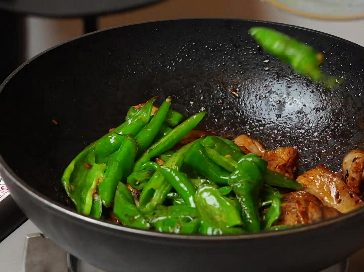 Eine Aufnahme aus der Vogelperspektive, die zeigt, wie gebräunte grüne Bratpaprika mit der gegarten Schweinefleischmischung in einem großen Wok vermengt werden.