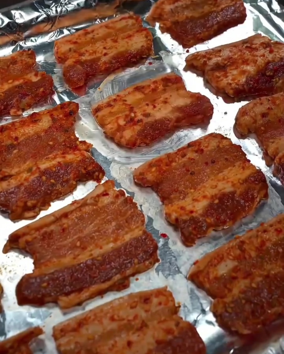 Speiseöl wird über die rohen Schweinebauchscheiben auf dem Backblech gesprüht.