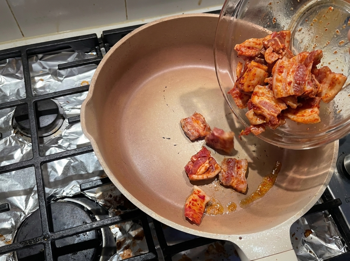 Marinierte Schweinebauchscheiben gleiten aus einer Glasschüssel in eine heiße Pfanne, um das Fett auszulassen.