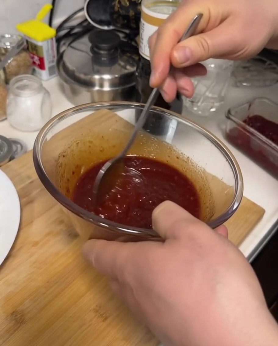Eine Hand rührt mit einem Metalllöffel eine dunkelrote, dicke Sauce in einer Glasschüssel um.