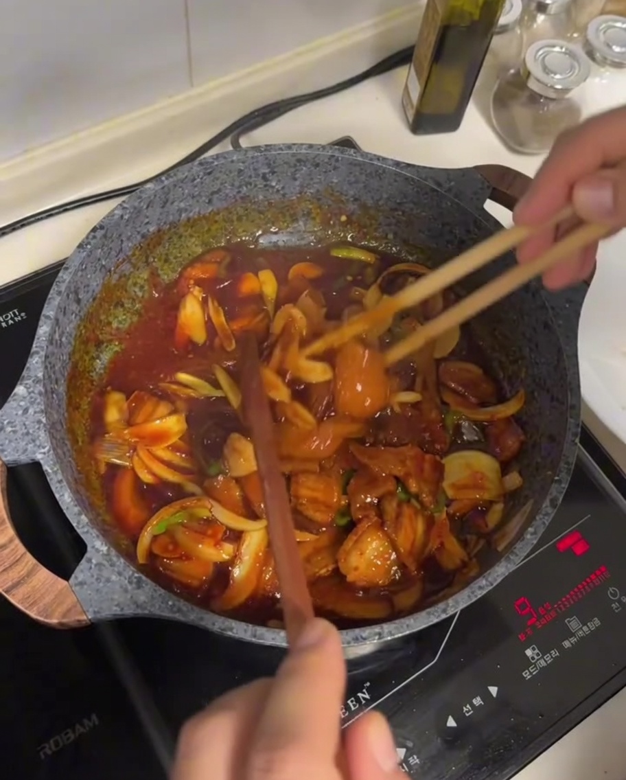 Schweinebauch und Gemüse werden in einer dunklen, glänzenden roten Sauce mit Stäbchen in einer Pfanne gewendet.