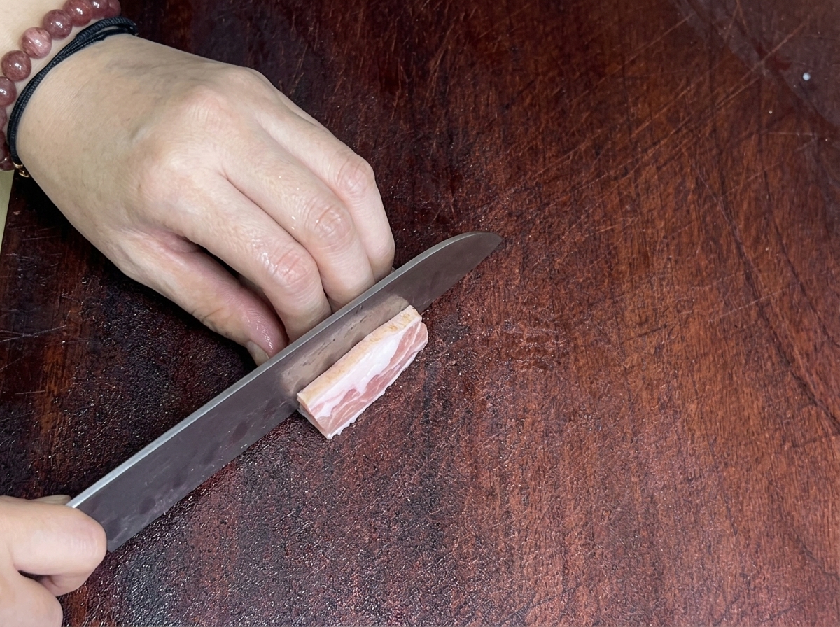 Nahaufnahme eines scharfen Messers beim Schneiden von Schweinebauch auf einem Holzbrett.