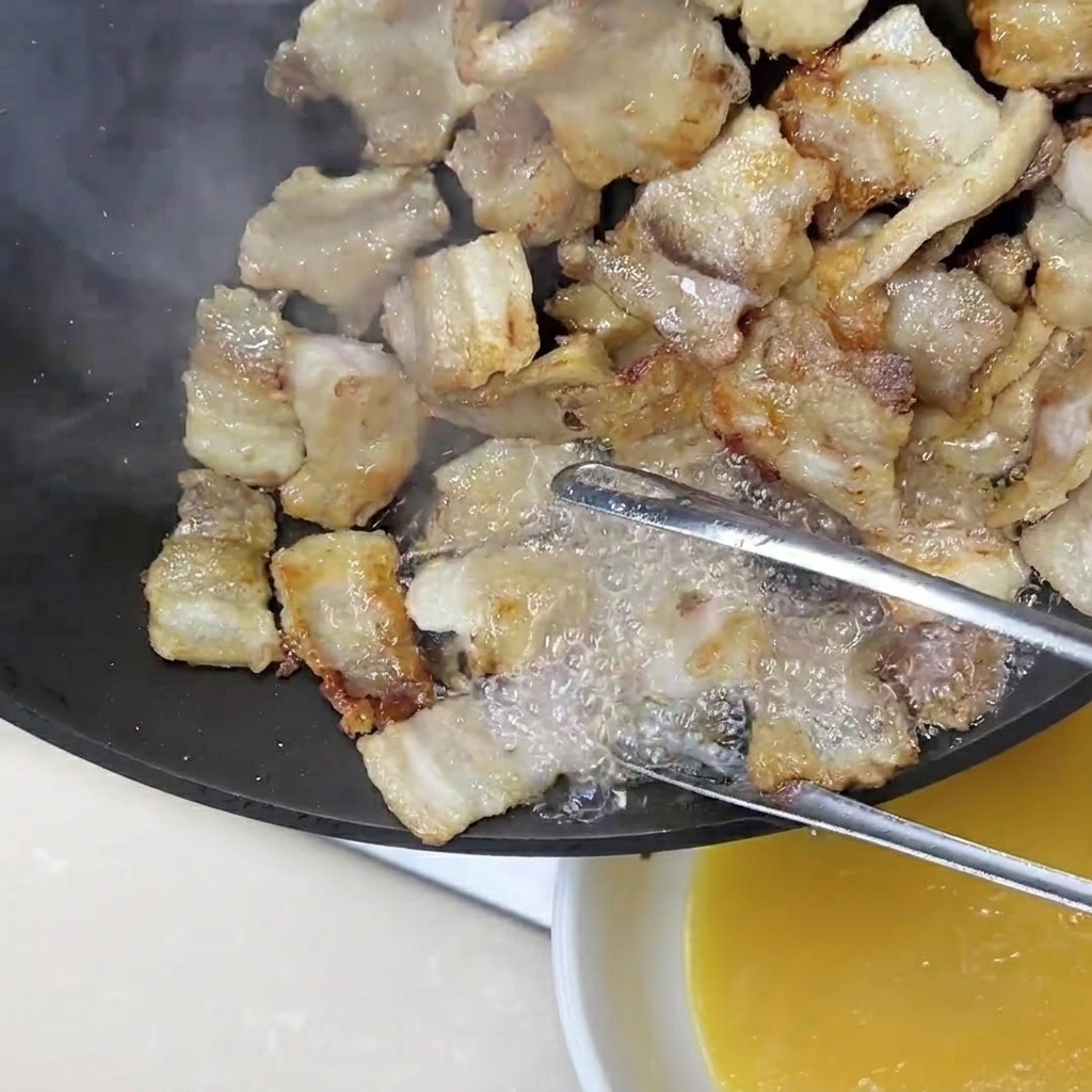 Eine Zange hält Schweinebauchscheiben, während goldenes Fett aus der Pfanne gegossen wird.
