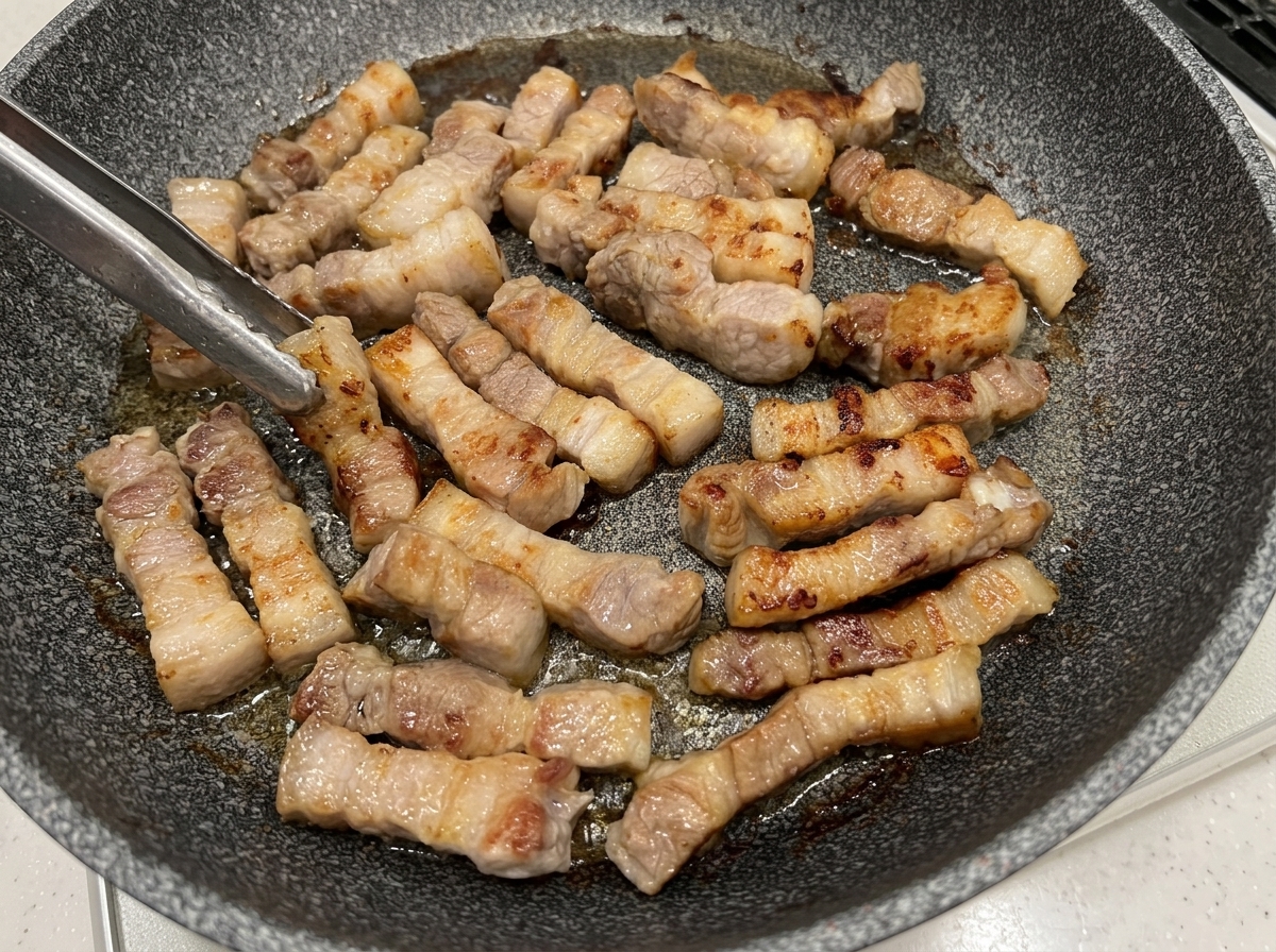 Schweinebauchstücke werden mit einer Zange in der Pfanne gewendet und zeigen goldbraune Oberflächen.