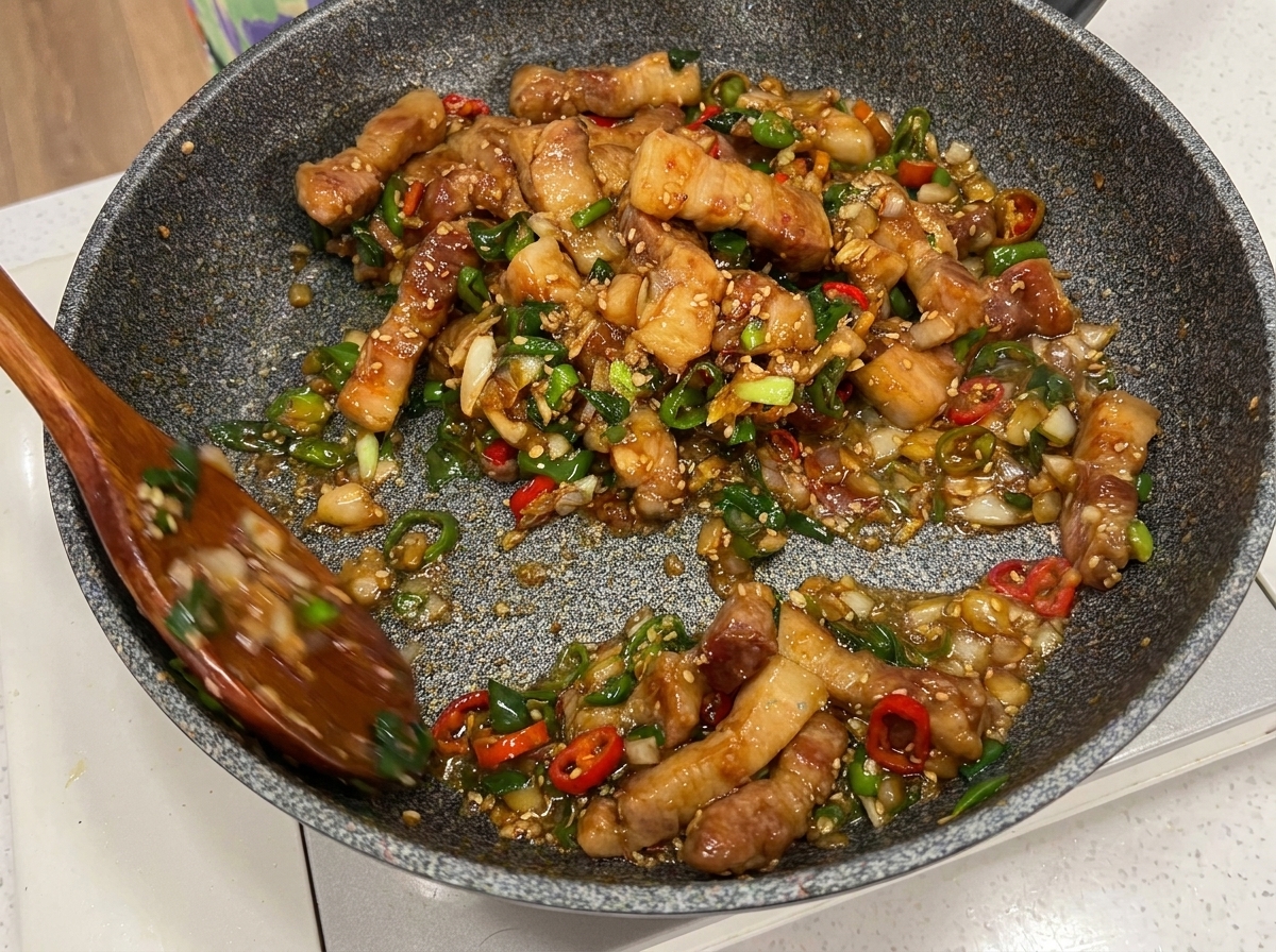 Schweinebauch und Gemüse werden mit zwei Holzlöffeln in der Pfanne vermengt.