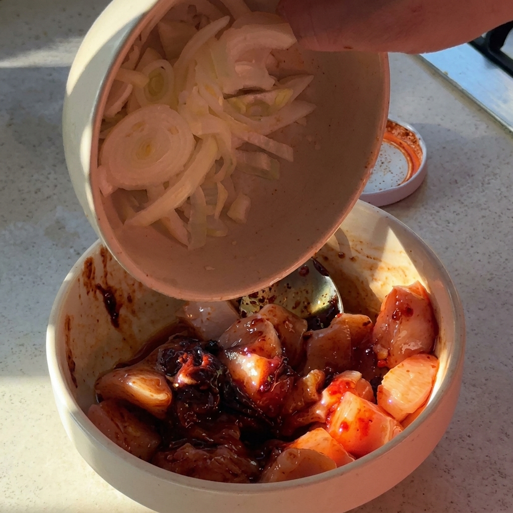 Geschnittene Zwiebeln und gehackter Knoblauch werden in eine Schüssel mit gewürzten Hähnchenstücken gegeben.