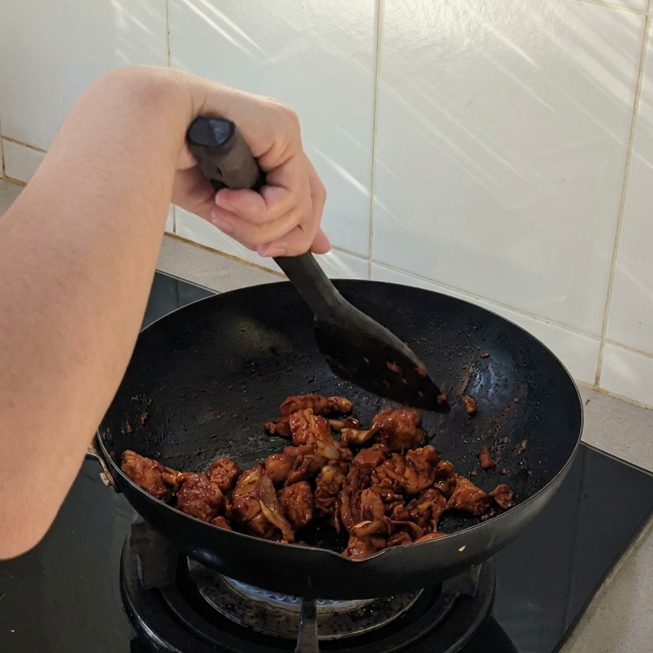 Hähnchen- und Zwiebelstücke werden in einem schwarzen Wok mit einem Pfannenwender gerührt, überzogen mit einer dunklen würzigen Sauce.