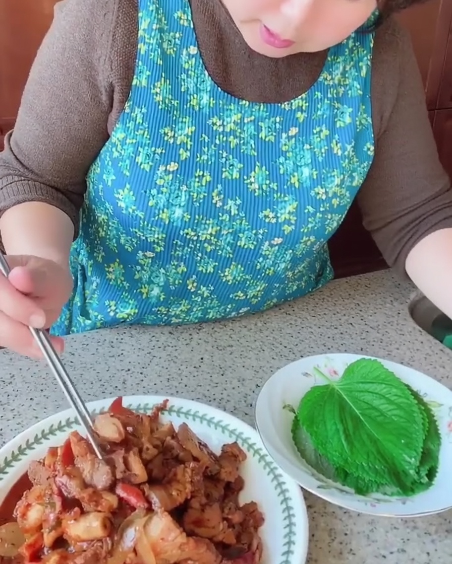 Eine Person legt mit Stäbchen ein Stück gekochten Schweinebauch und Tintenfisch auf ein frisches grünes Perilla-Blatt.