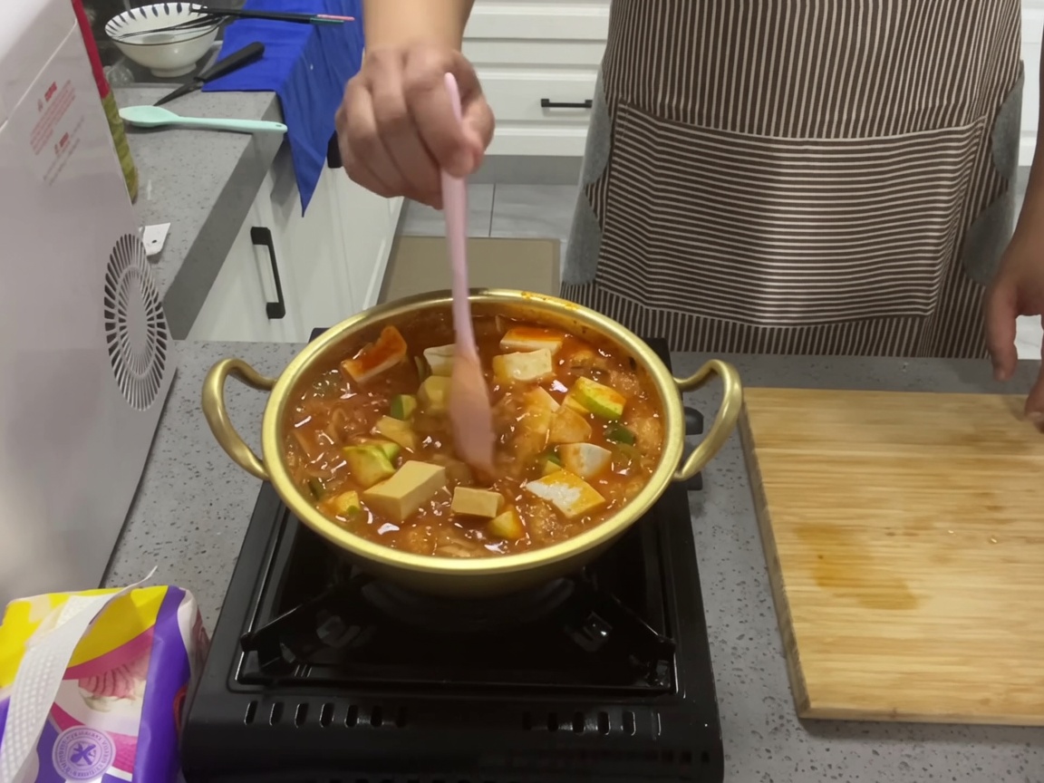Eine Nahaufnahme eines blubbernden Thunfisch- und Kimchi-Eintopfs in einem goldfarbenen Topf, der mit einem rosa Silikonlöffel umgerührt wird, um Tofu und Zucchini zu vermischen.