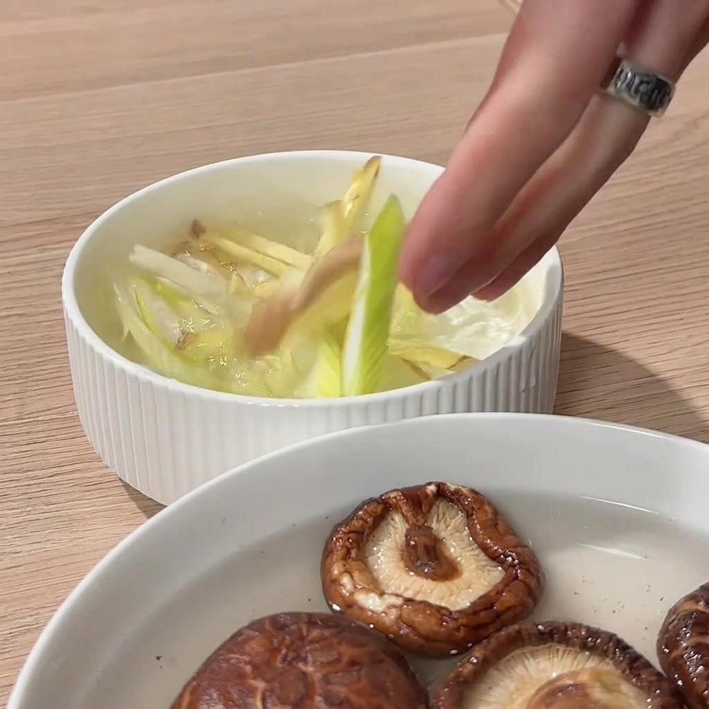 Eine Hand drückt Ingwerscheiben und Frühlingszwiebeln in einer Schüssel mit Wasser, um einen aromatischen Aufguss zu erzeugen.