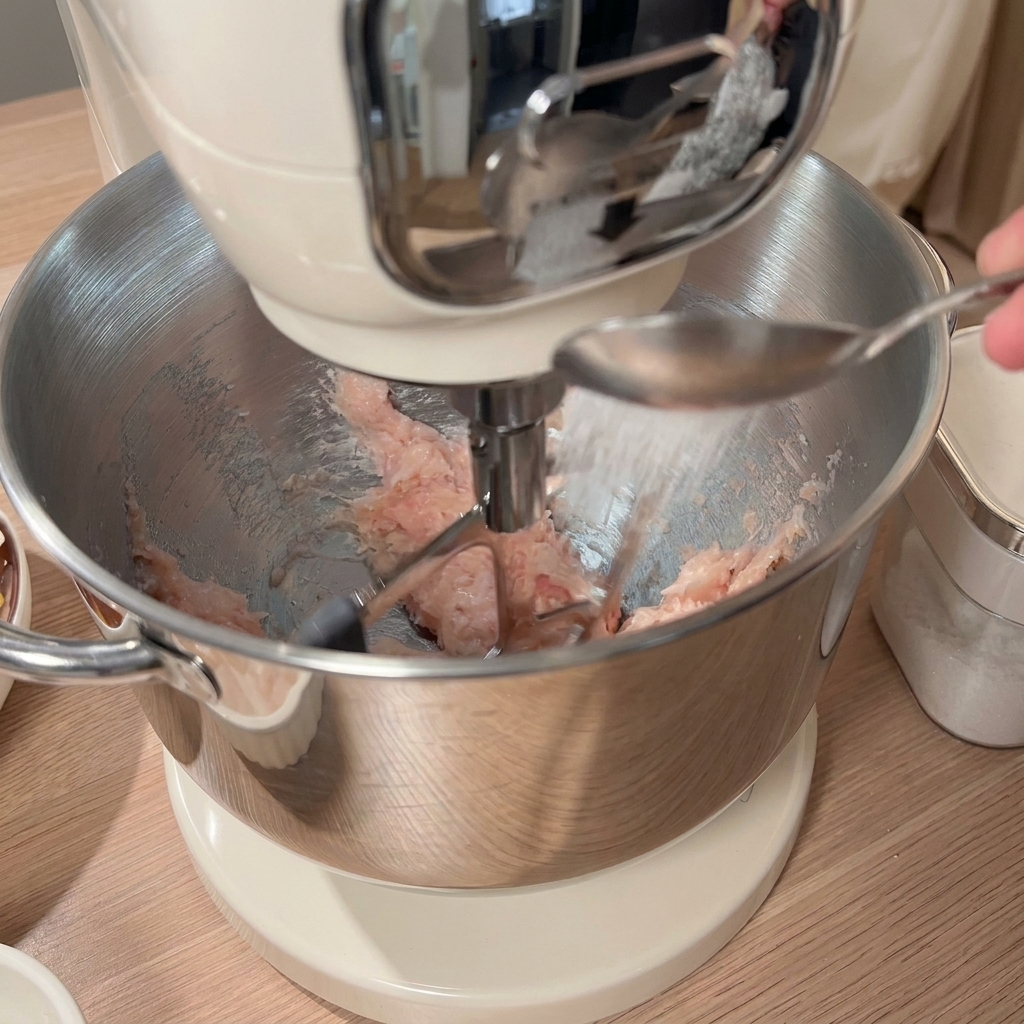 Ein Löffel mit Gewürzen wird zum gehackten Huhn in die Rührschüssel gegeben, wobei weitere Zutaten auf dem Tisch sichtbar sind.