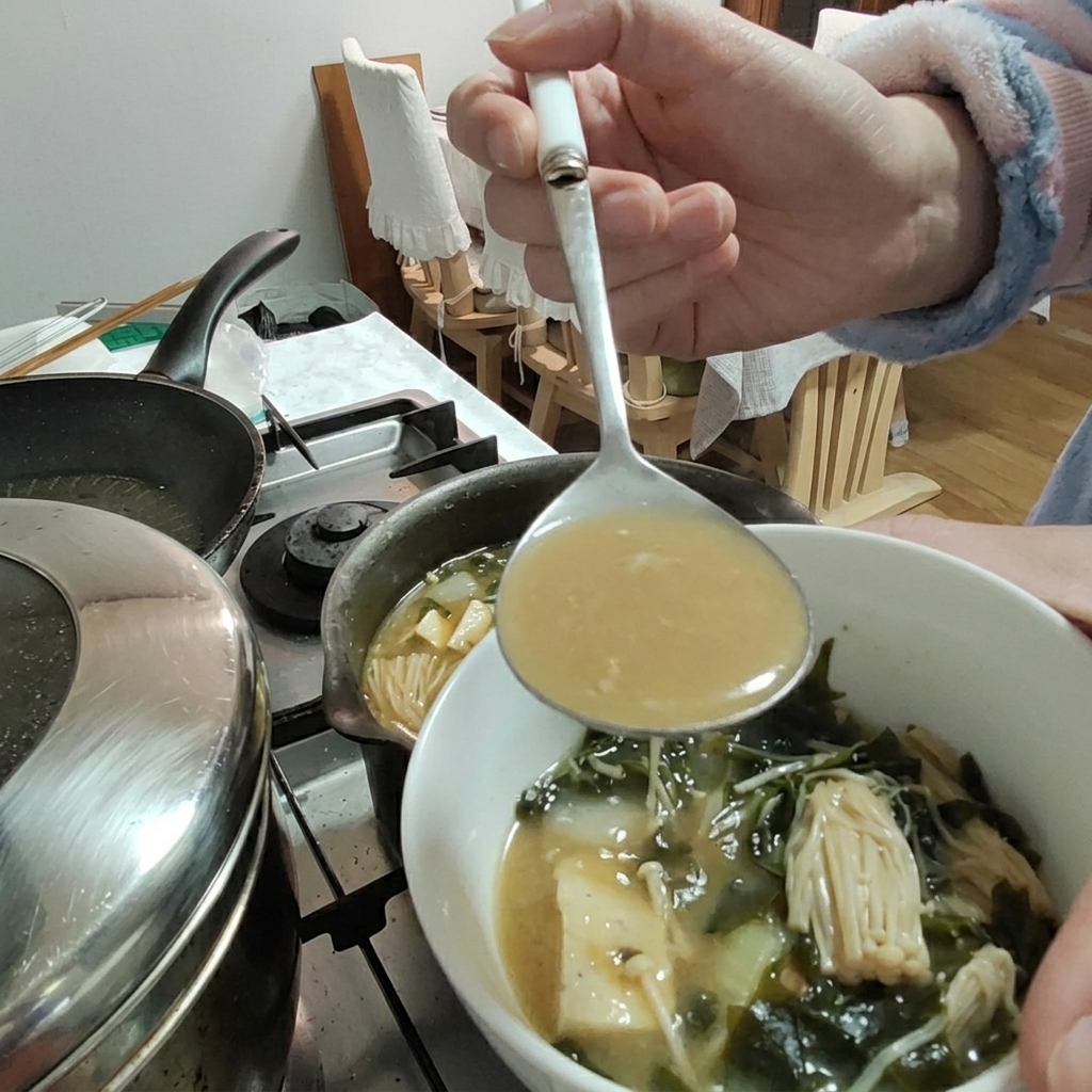 Sojabohnenpaste wird mit einer Kelle in den Eintopf gegeben, Tofu und Enoki-Pilze sind in der Schüssel sichtbar.
