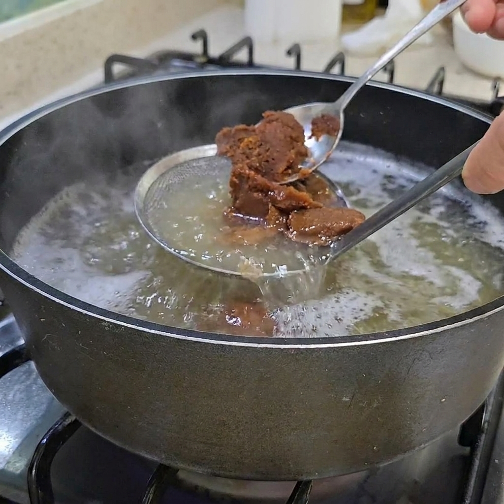 Zwei Löffel dicker, brauner koreanischer Sojabohnenpaste werden in einen Topf mit klarer Brühe gerührt.