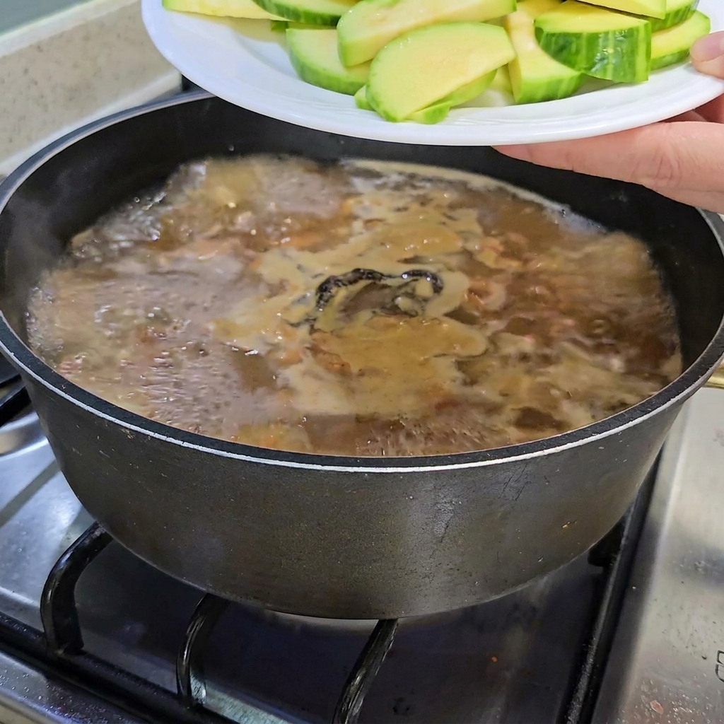 Geschnittene Zucchini und ein Bund Enoki-Pilze werden in den köchelnden Eintopftopf gegeben.