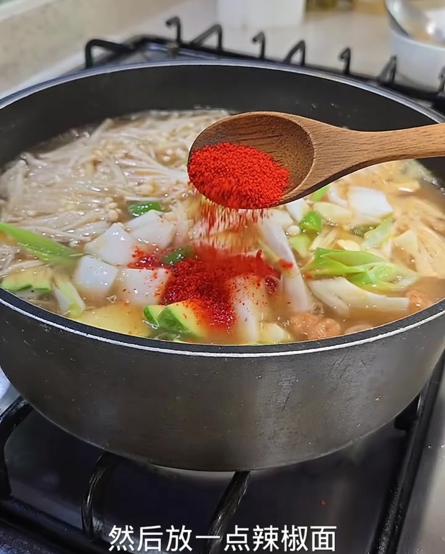 Eine Nahaufnahme von leuchtend rotem Chilipulver, das von einem Holzlöffel in den köchelnden Eintopf mit Zucchini, Zwiebeln und Enoki-Pilzen gestreut wird.