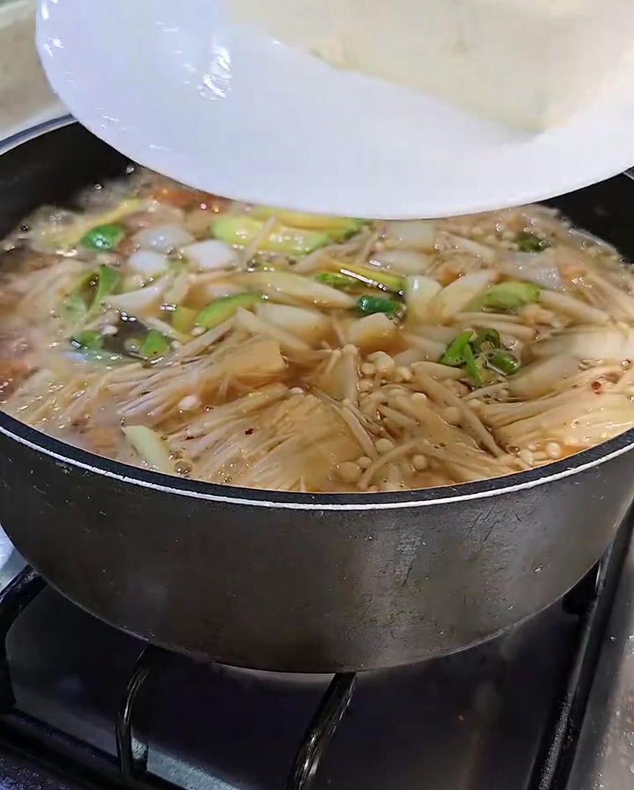 Scheiben von weißem Tofu werden vorsichtig von einem weißen Teller in den blubbernden, aromatischen Sojabohnenpasten-Eintopf gegeben.