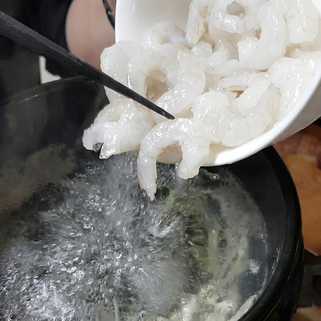 Eine weiße Schüssel gibt frische Garnelen in einen blubbernden Steintopf.