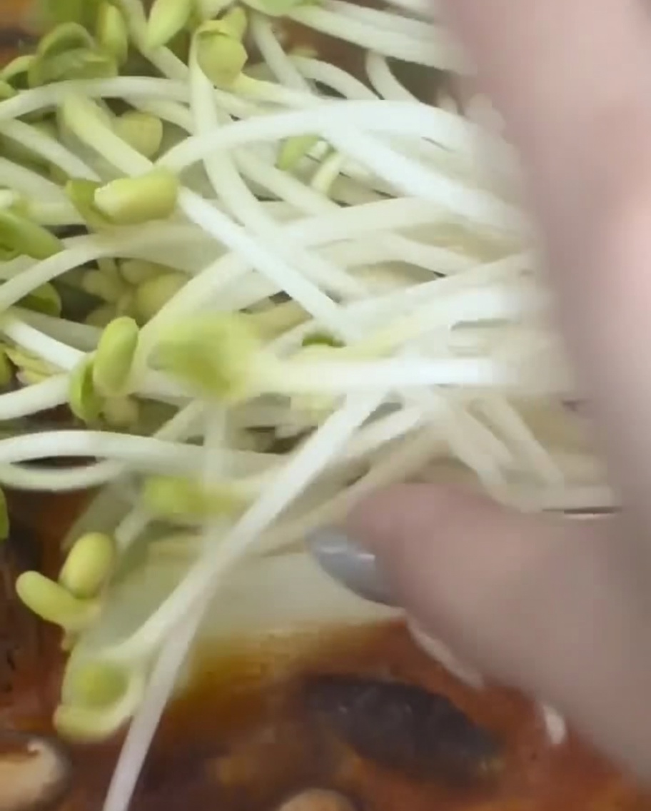 Eine Handvoll frischer Sojabohnensprossen wird in einen Topf mit köchelndem koreanischem Eintopf gegeben.