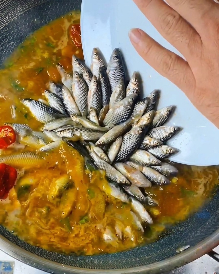 Eine Hand gießt flüssige Würze aus einer Flasche über die angeordneten Fische im Wok.