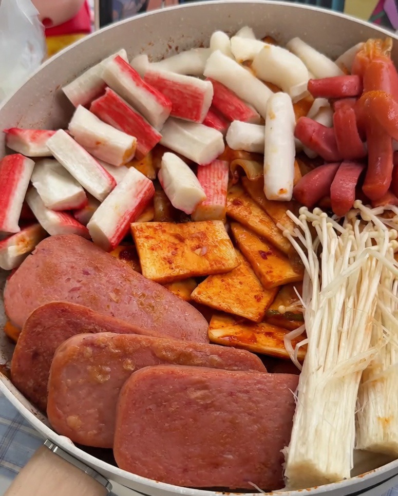 Eine Pfanne, wunderschön arrangiert mit ordentlichen Bereichen aus in Scheiben geschnittenem Spam, Surimi, Würstchen, Reiskuchen, Fischkuchen und Enoki-Pilzen.