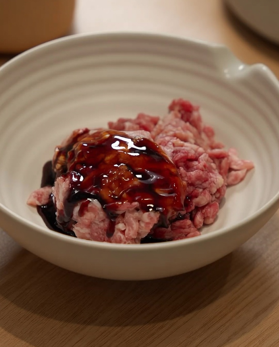 Schweinehackfleisch in einer weißen Schüssel, das mit Sojasauce und Kochwein gewürzt wird.