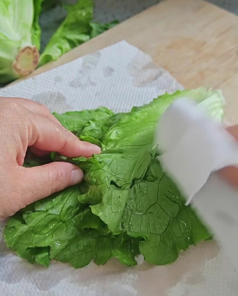 Hände trocknen frische, grüne Salatblätter mit weißen Papiertüchern auf einem Holzbrett ab.