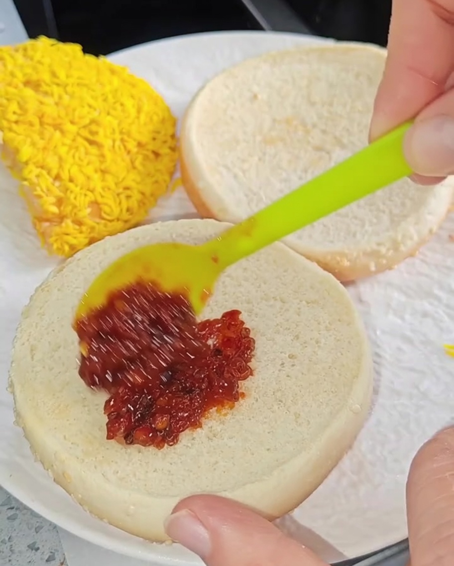 Ein kleiner Spatel verstreicht rote Chilisauce auf der Innenseite eines gerösteten Burger-Brötchens.