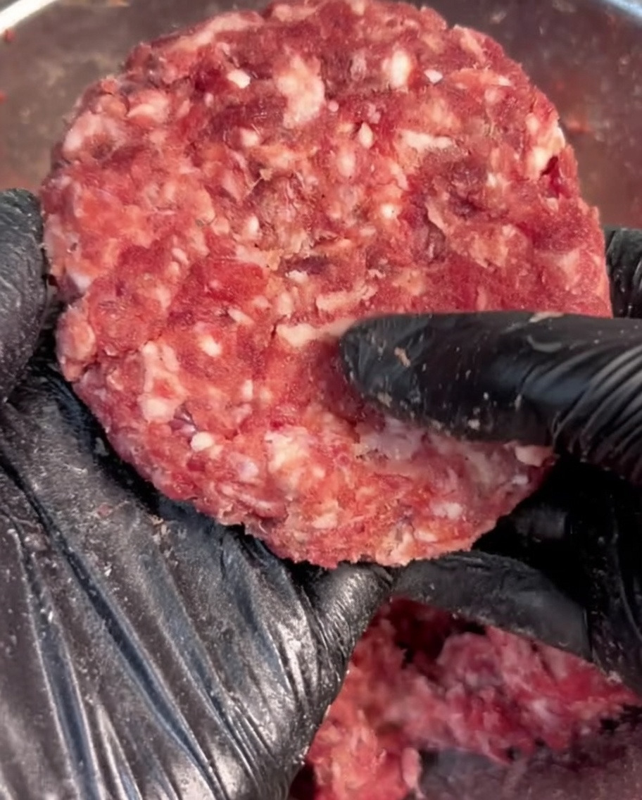 Eine Hand in einem schwarzen Handschuh formt gewürztes Rinderhackfleisch zu einem runden Patty.