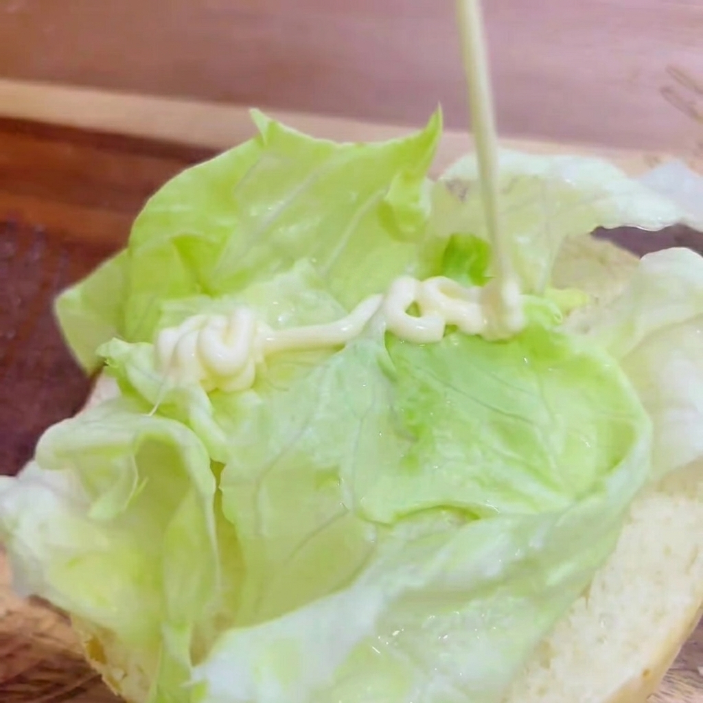 Mayonnaise wird in Zickzacklinien auf den Salat auf dem Burgerbrötchen gespritzt.