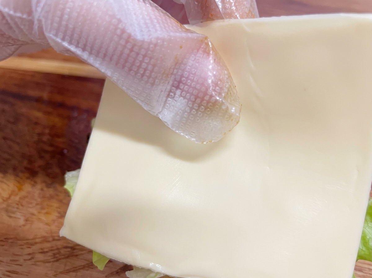 Eine behandschuhte Hand legt eine Scheibe Käse auf den Burger.