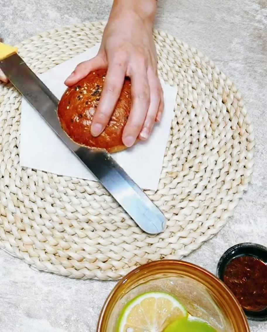 Eine Hand hält ein Messer und schneidet ein weiches, hausgemachtes Burgerbrötchen auf einem Holzbrett durch.