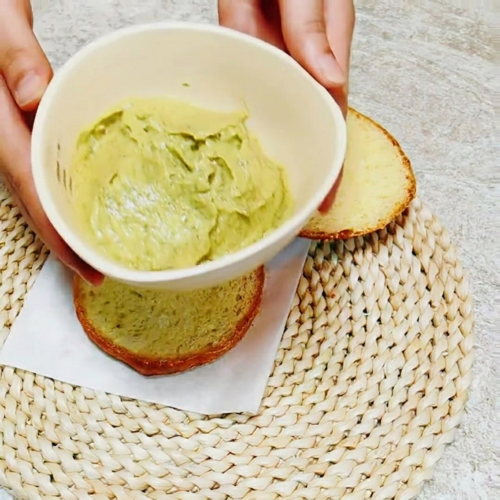 Nahaufnahme: Ein Messer verteilt die hellgrüne Avocado-Basilikum-Creme auf der Innenseite eines Brötchens.