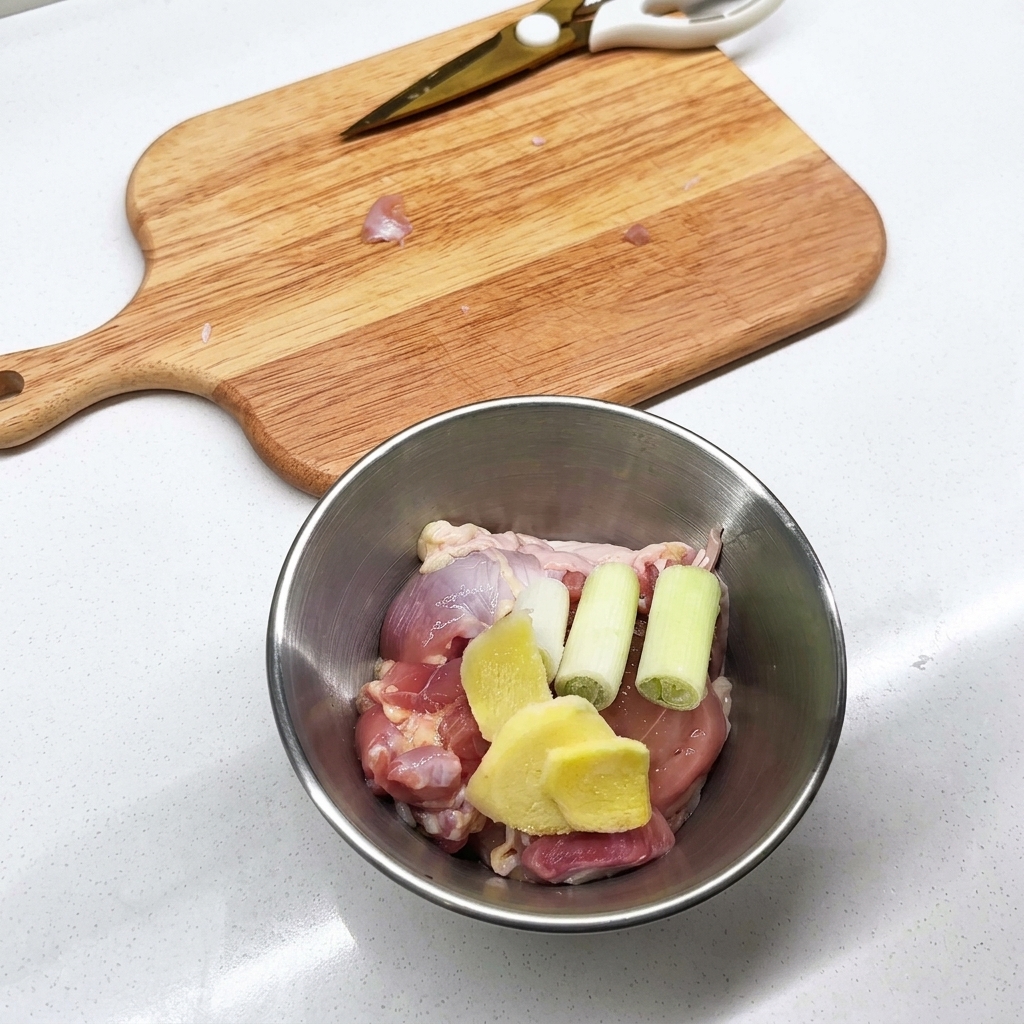 Metallschüssel mit rohen Hähnchenstücken, geschnittenem Ingwer und Frühlingszwiebeln.