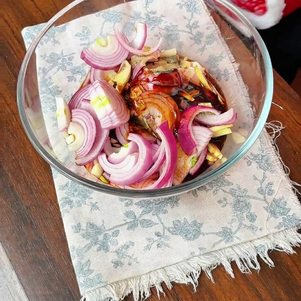 Hähnchenschenkel, Zwiebelscheiben und dunkle Marinadengewürze vermischt in einer großen Glasschüssel.