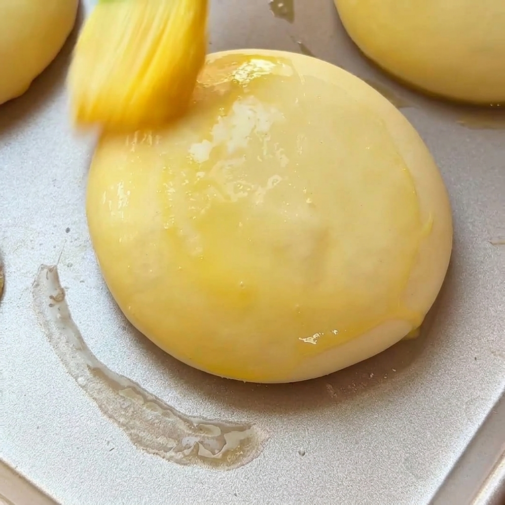 Ein Backpinsel bestreicht ein aufgegangenes Burgerbrötchen auf einem Backblech mit Eistreiche.