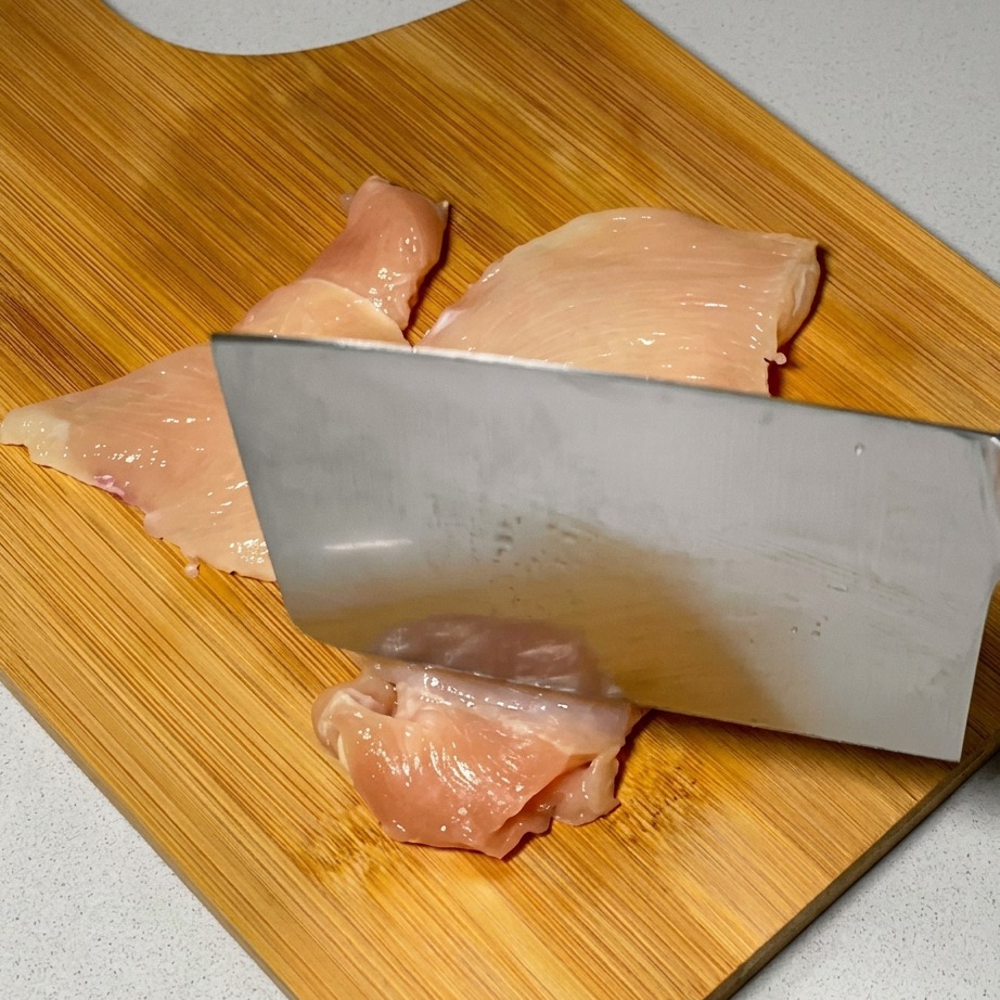 Ein Küchenmesser wird verwendet, um rohe Hähnchenscheiben auf einer Holzoberfläche zartzuklopfen.