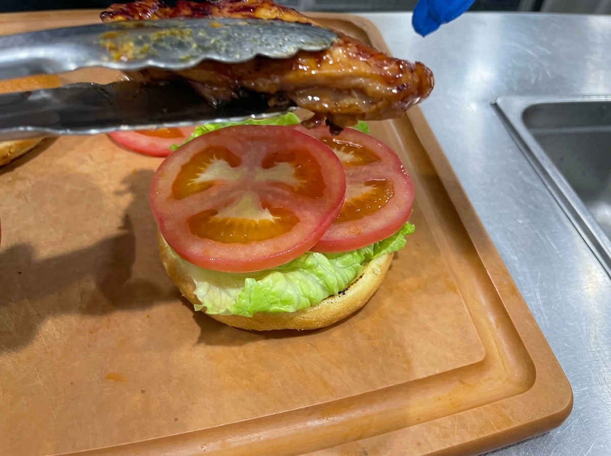 Ein gebratener Teriyaki-Hähnchenschenkel wird mit einer Zange auf das mit Salat und Tomatenscheiben belegte Burgerbrötchen gelegt.