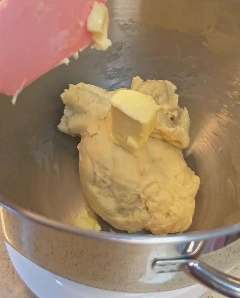 Ein Stück Butter wird dem bereits teilweise gekneteten Teig in der Edelstahl-Rührschüssel hinzugefügt.