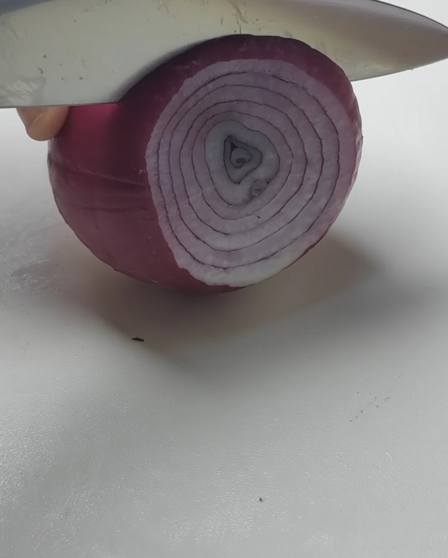 Ein Messer schneidet eine rote Zwiebel in dicke Ringe auf einem weißen Schneidebrett.