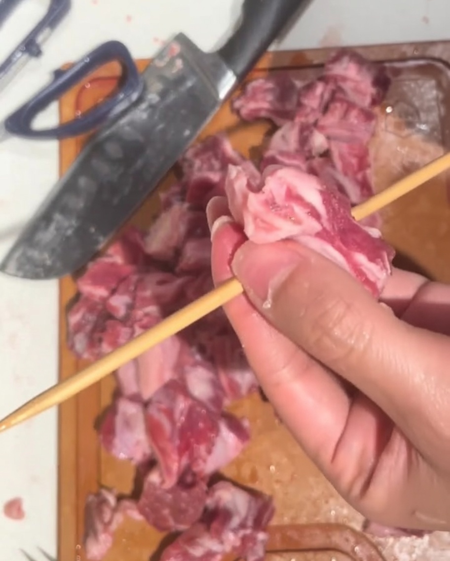 Eine Hand schiebt ein gewürfeltes Stück rohes Rindfleisch auf einen langen Holzspieß.