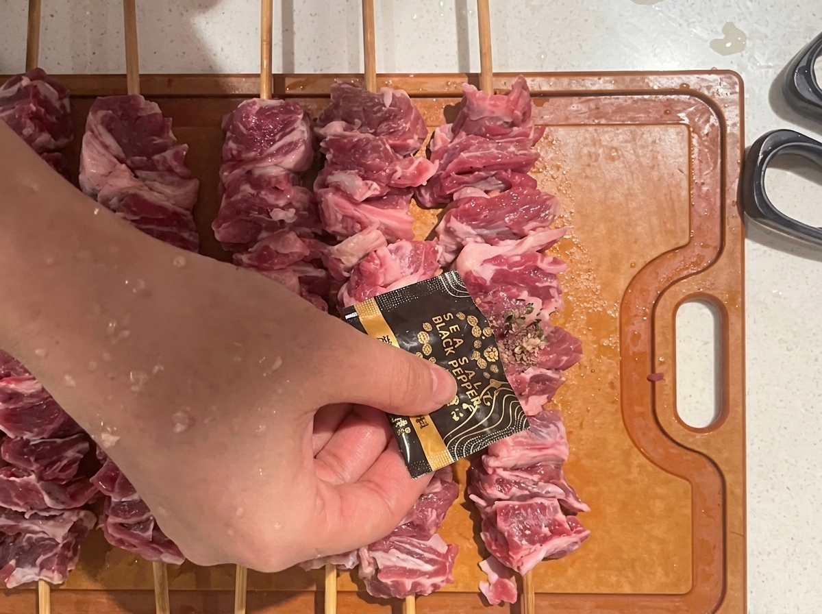 Eine Hand streut Gewürze über rohe Rindfleischspieße auf einem Holzbrett.