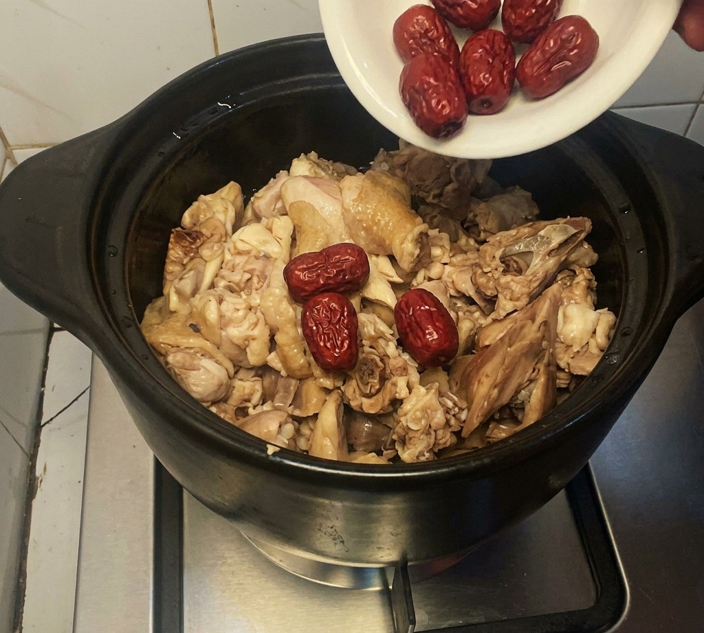 Ein kleiner weißer Teller gibt getrocknete rote Datteln in einen Tontopf mit halb gegartem Huhn.