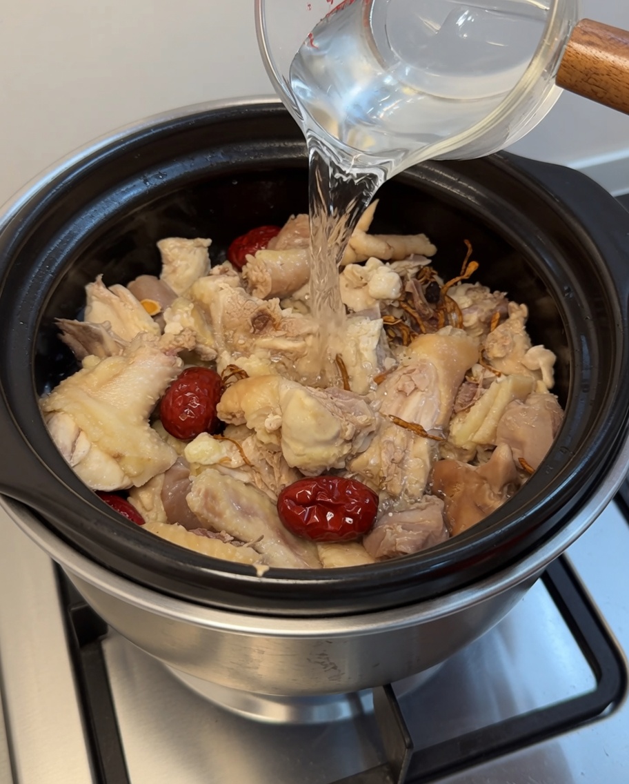Klares Wasser wird aus einem Messbecher in einen Tontopf mit Huhn, Datteln und Kräutern gegossen.