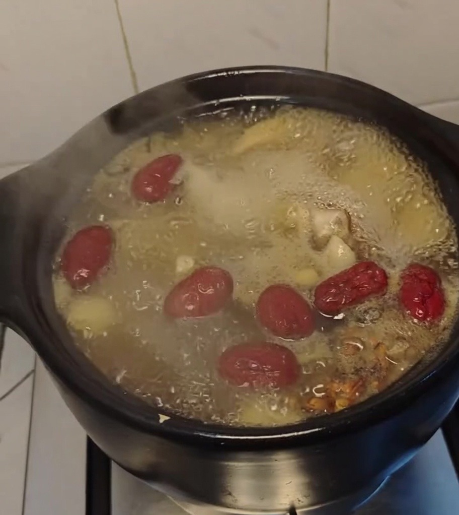 Hühnersuppe köchelt in einem schwarzen Tontopf, große rote Datteln schwimmen an der Oberfläche.