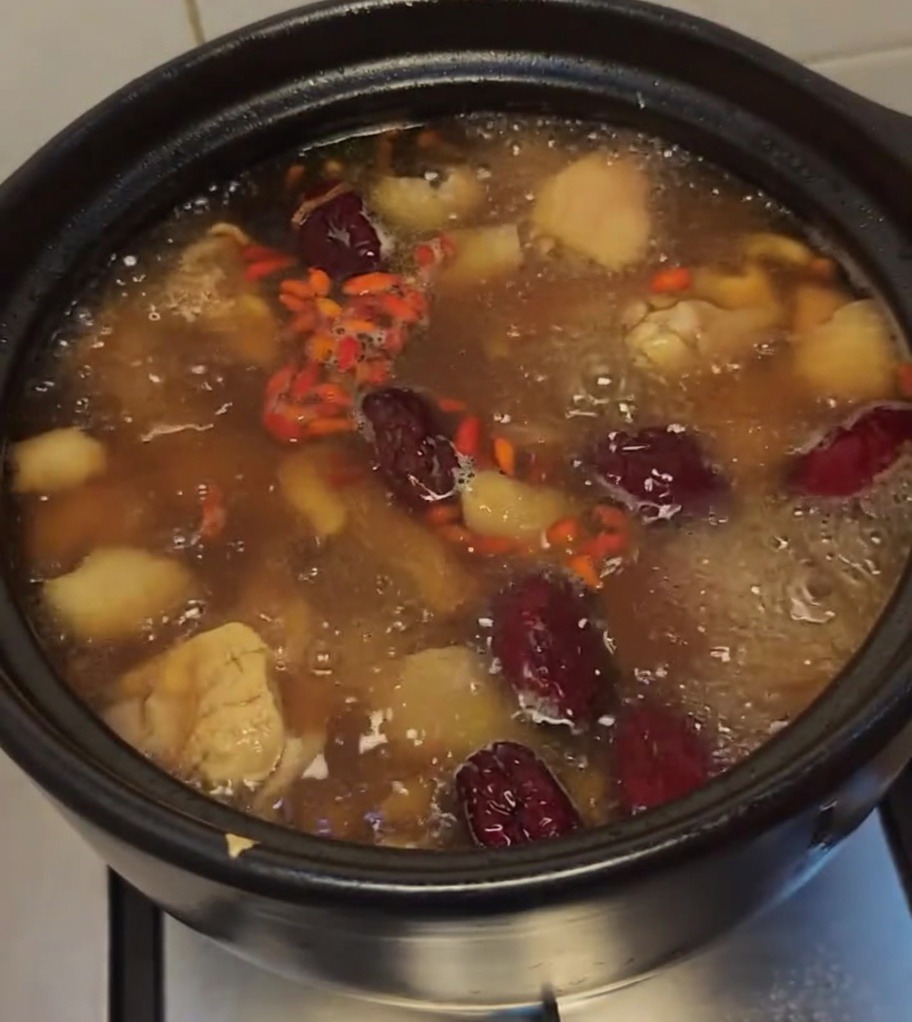 Kochende Hühnersuppe im schwarzen Tontopf, garniert mit leuchtend roten Goji-Beeren und roten Datteln.