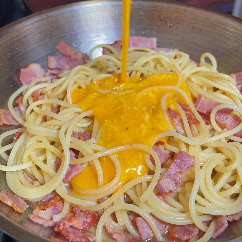 Die gelbe Ei-Käse-Soße wird über die heißen Nudeln und den Speck in die Pfanne gegossen.