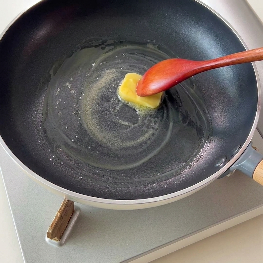 Ein Stück gelbe Butter schmilzt in einer Pfanne und wird mit einem Holzlöffel gerührt.