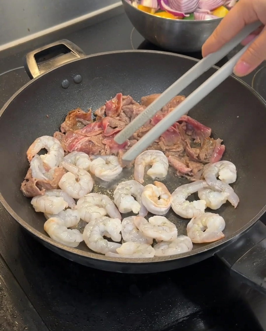 Rindfleischscheiben und rohe graue Garnelen werden zusammen in einem heißen Wok gebraten.